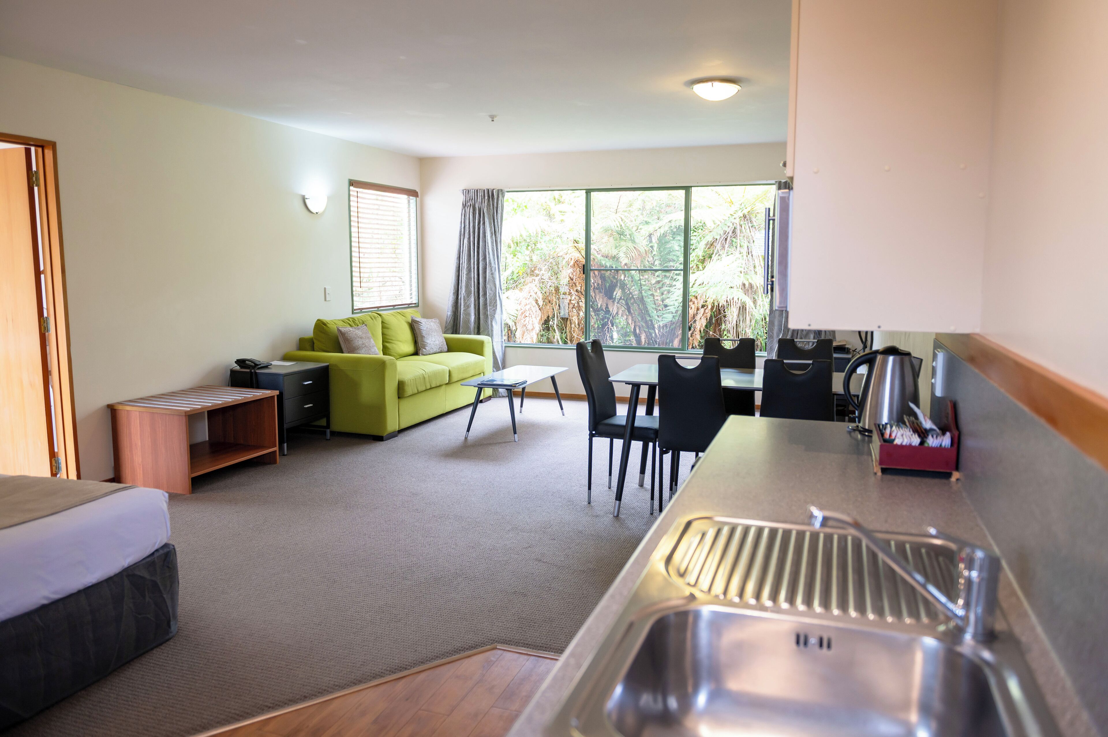 Una habitación de hotel moderna con sofá, una mesa de comedor, sillas, una pequeña cocina con fregadero y un amplio ventanal con vistas a zonas verdes.