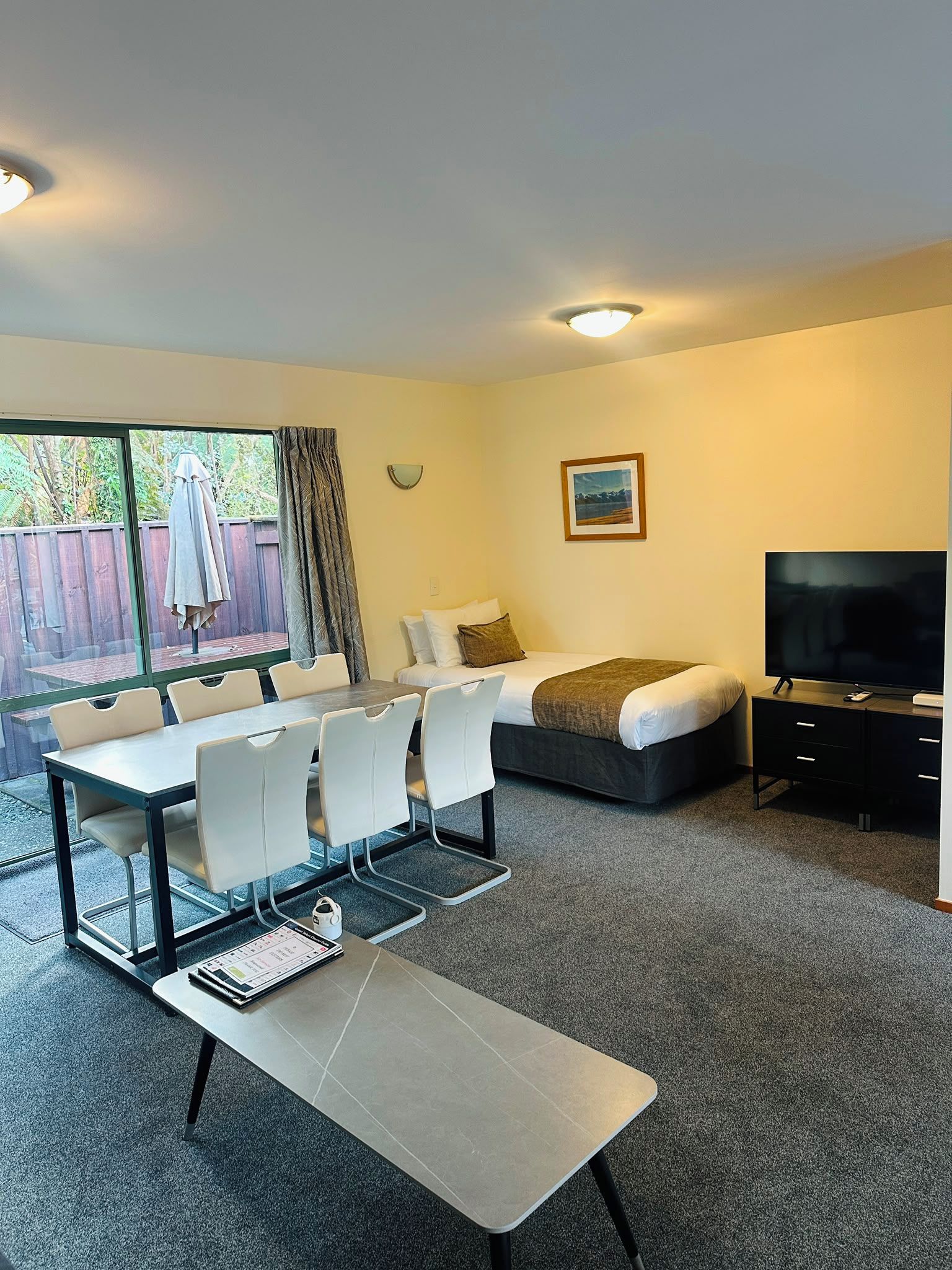 A room with a bed, a TV, a dining table with chairs, and a window with curtains.