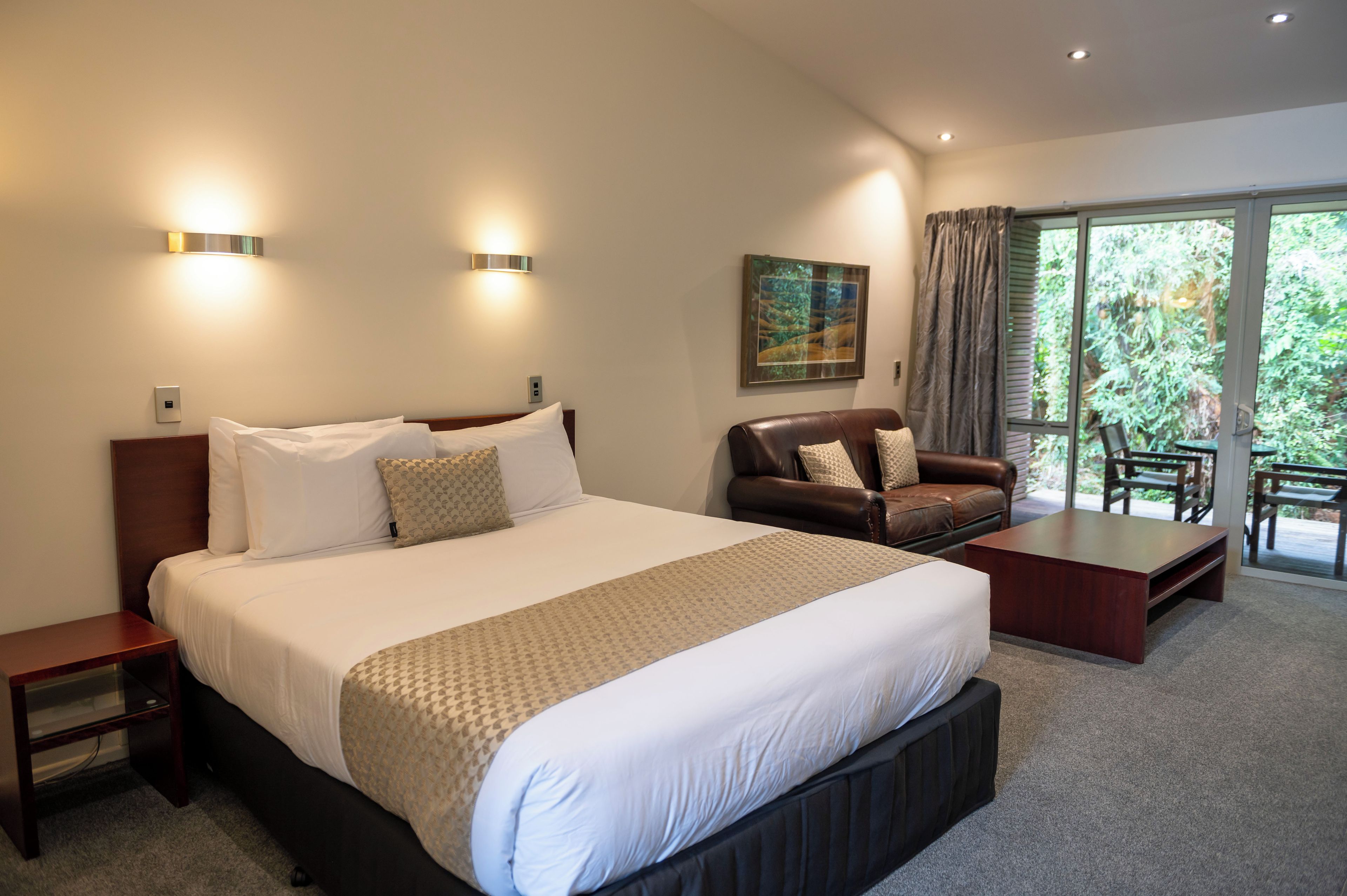 Une chambre d’hôtel avec un grand lit, un canapé en cuir, une table basse et un balcon donnant sur un paysage verdoyant.