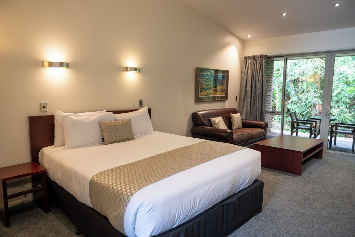 Une chambre d’hôtel avec un grand lit, un canapé en cuir, une table basse et un balcon donnant sur un paysage verdoyant.