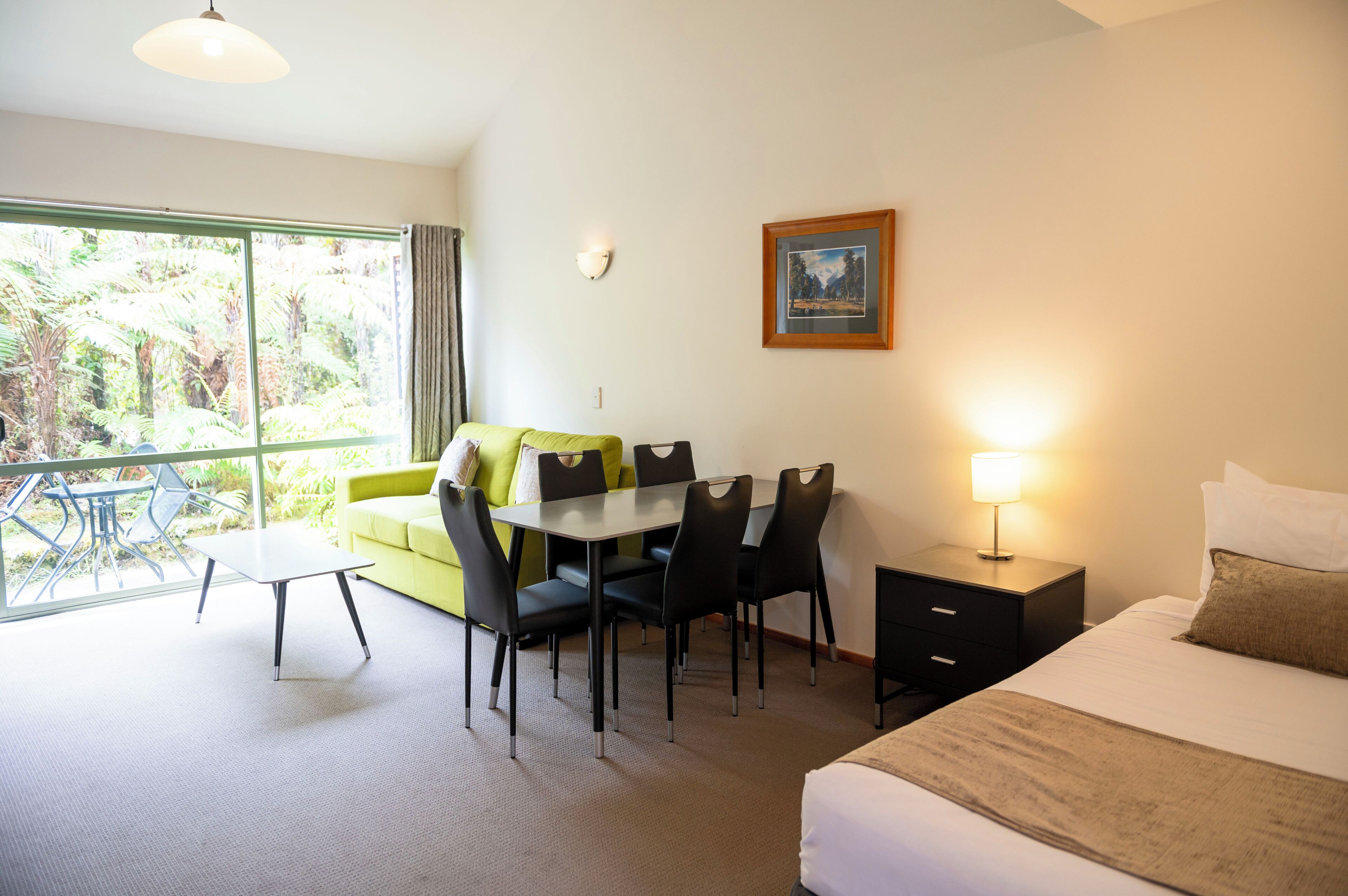Habitación de hotel con cama, una mesa de comedor con sillas, una mesita, una mesita de noche con lámpara, un cuadro en la pared y un ventanal con vistas a la vegetación.