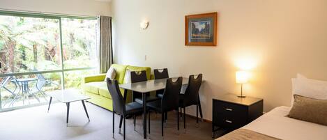 Habitación de hotel con cama, una mesa de comedor con sillas, una mesita, una mesita de noche con lámpara, un cuadro en la pared y un ventanal con vistas a la vegetación.