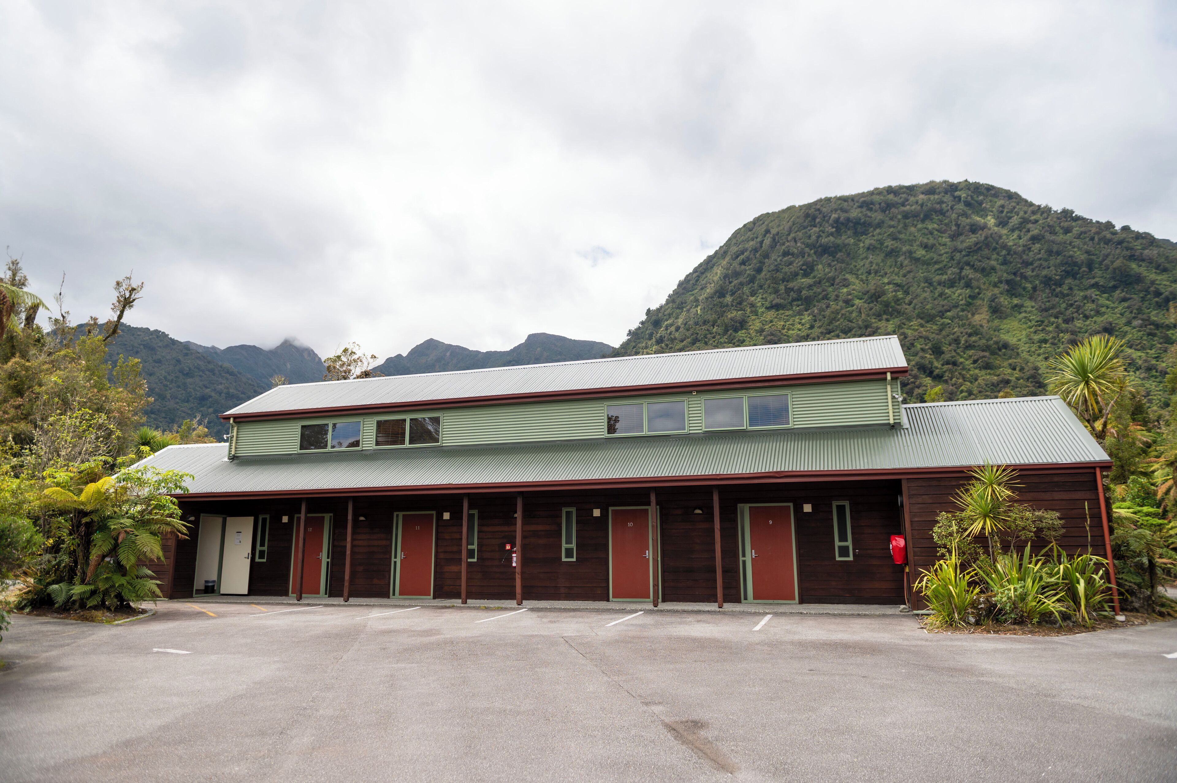 A two-story building with a green roof and multiple doors, surrounded by lush greenery and mountains in the background.