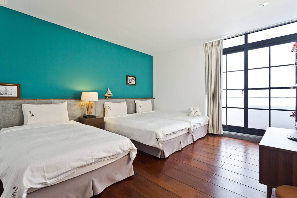 Quarto de hotel com duas camas, uma escrivaninha, uma janela com cortinas e parede cor de verde-água.