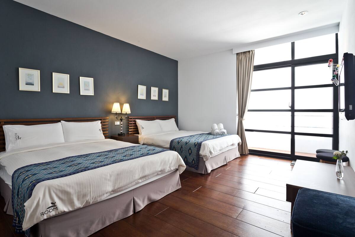 Quarto de hotel com duas camas, uma janela grande e uma mesa com um vaso.