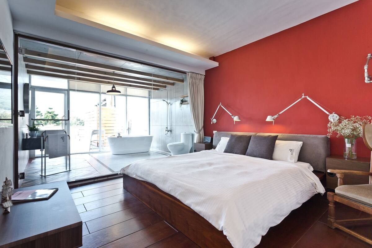 Quarto moderno com uma cama grande, uma parede de destaque vermelha e vista para o exterior.