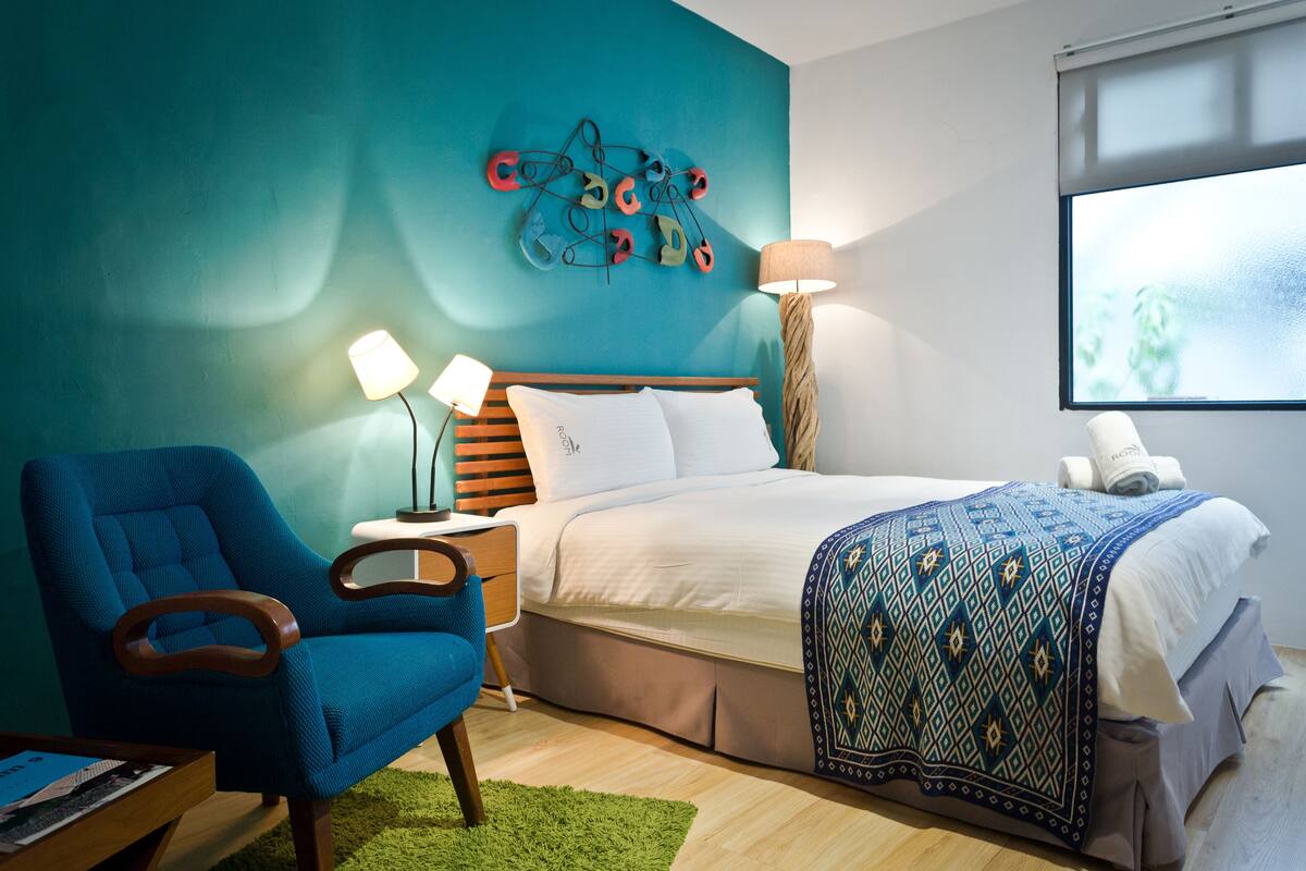 Quarto de hotel com uma cama, uma poltrona azul, uma mesa de cabeceira com luminárias e uma obra de arte na parede.