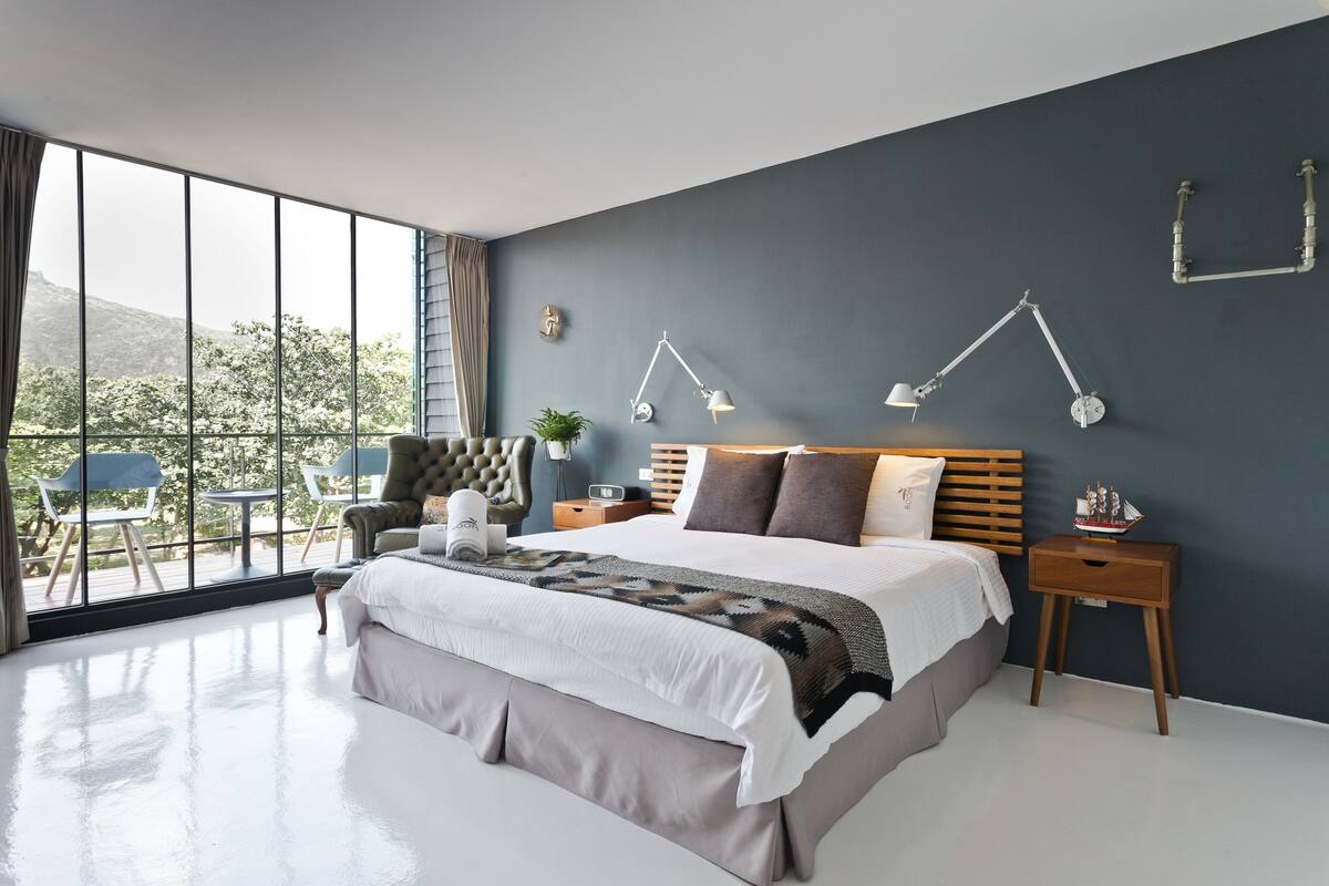 Quarto moderno com cama grande, mesas de cabeceira, um criado-mudo e vista para o exterior através de amplas janelas.