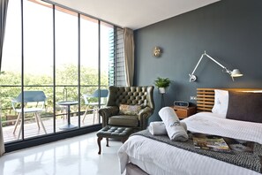 A modern hotel room with a large window, a comfortable armchair, a neatly made bed, and a bedside table with a lamp.