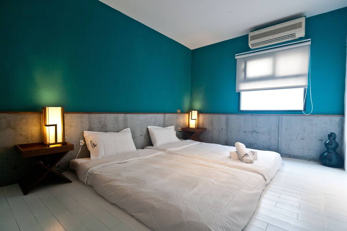 Quarto com paredes verde-água, uma cama grande com roupa de cama branca, duas luminárias de cabeceira e um aparelho de ar condicionado sobre uma janela.