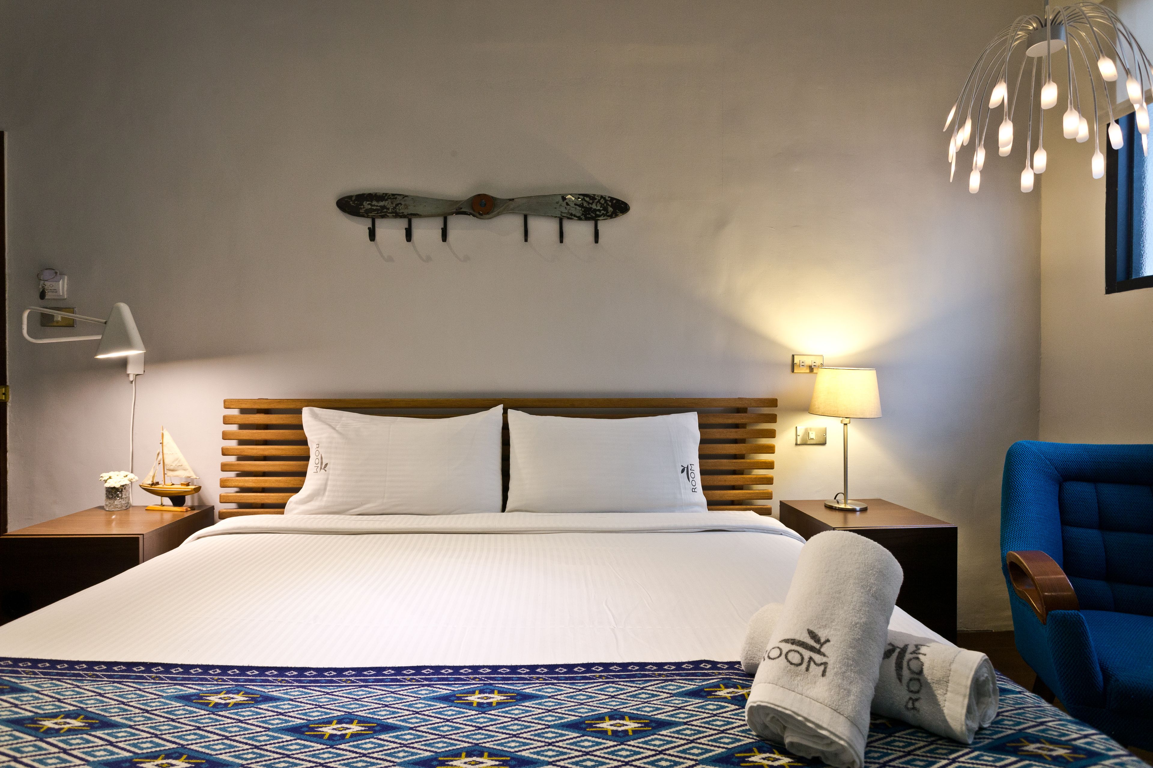 Habitación de hotel con una cama grande, cabecera de madera, dos lámparas de noche, un adorno en la pared, un sillón azul y una alfombra estampada.