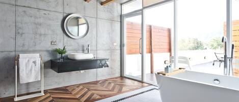 Un baño moderno con un ventanal grande, una bañera independiente, un espejo redondo y suelo de madera.