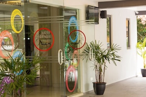 Entrada de un hotel con puertas de vidrio, plantas en macetas y varios adhesivos circulares en el cristal.