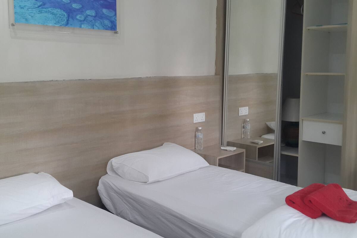 A hotel room with two single beds, a small bedside table with a water bottle and remote control, a mirror, and a wardrobe with a hanging rack.