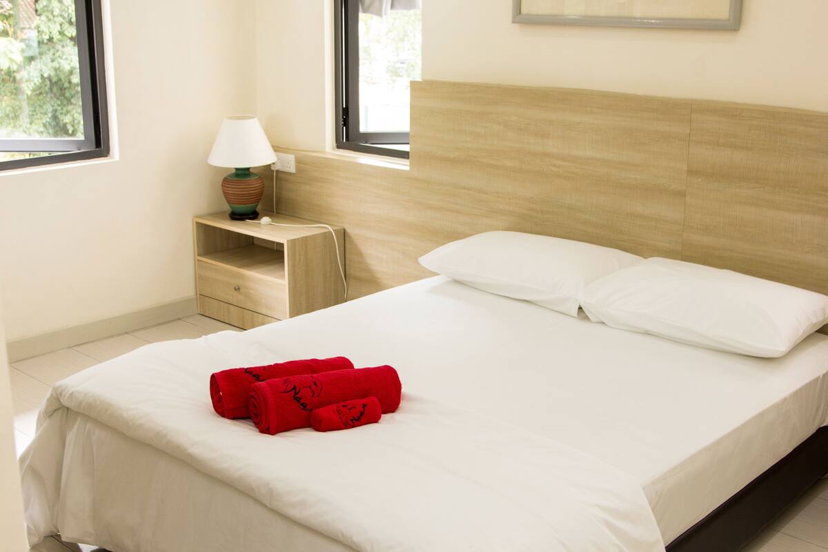 A neatly made bed with white linens and red towels, a bedside lamp, and a window with a view of greenery.