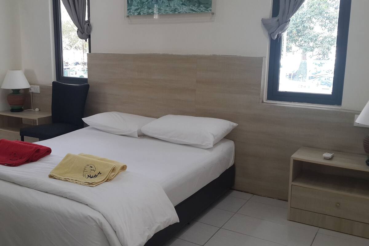 A neatly made bed with white linens and two pillows, a red towel, and a yellow towel on the bed. There is a nightstand with a lamp and a small table with a lamp on it. The room has a window with curtains, a wall-mounted painting, and a tiled floor.