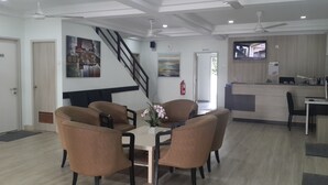 A modern dining area with a wooden table and four brown chairs, a staircase, and a reception desk with a TV. - Naam Boutique Lodge (Port Dickson)