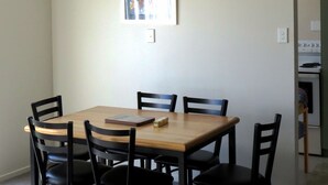 Un coin repas avec une table en bois et six chaises, un tableau encadré au mur, et une cuisine visible au fond.