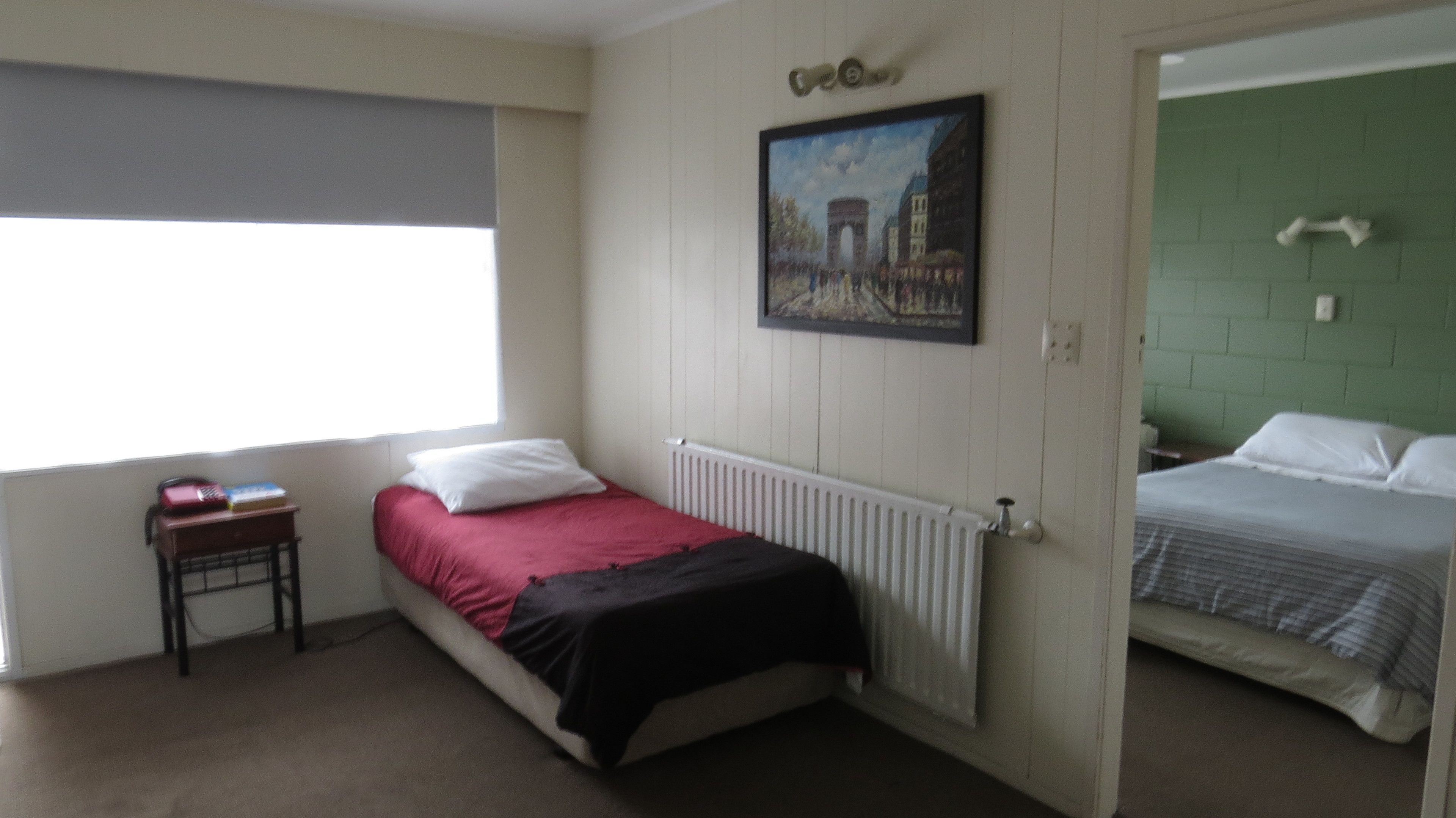 A room with two beds, one with a red and black bedspread and another with a striped bedspread. There is a radiator under the first bed, a small table with a book and a lamp, and a painting on the wall.