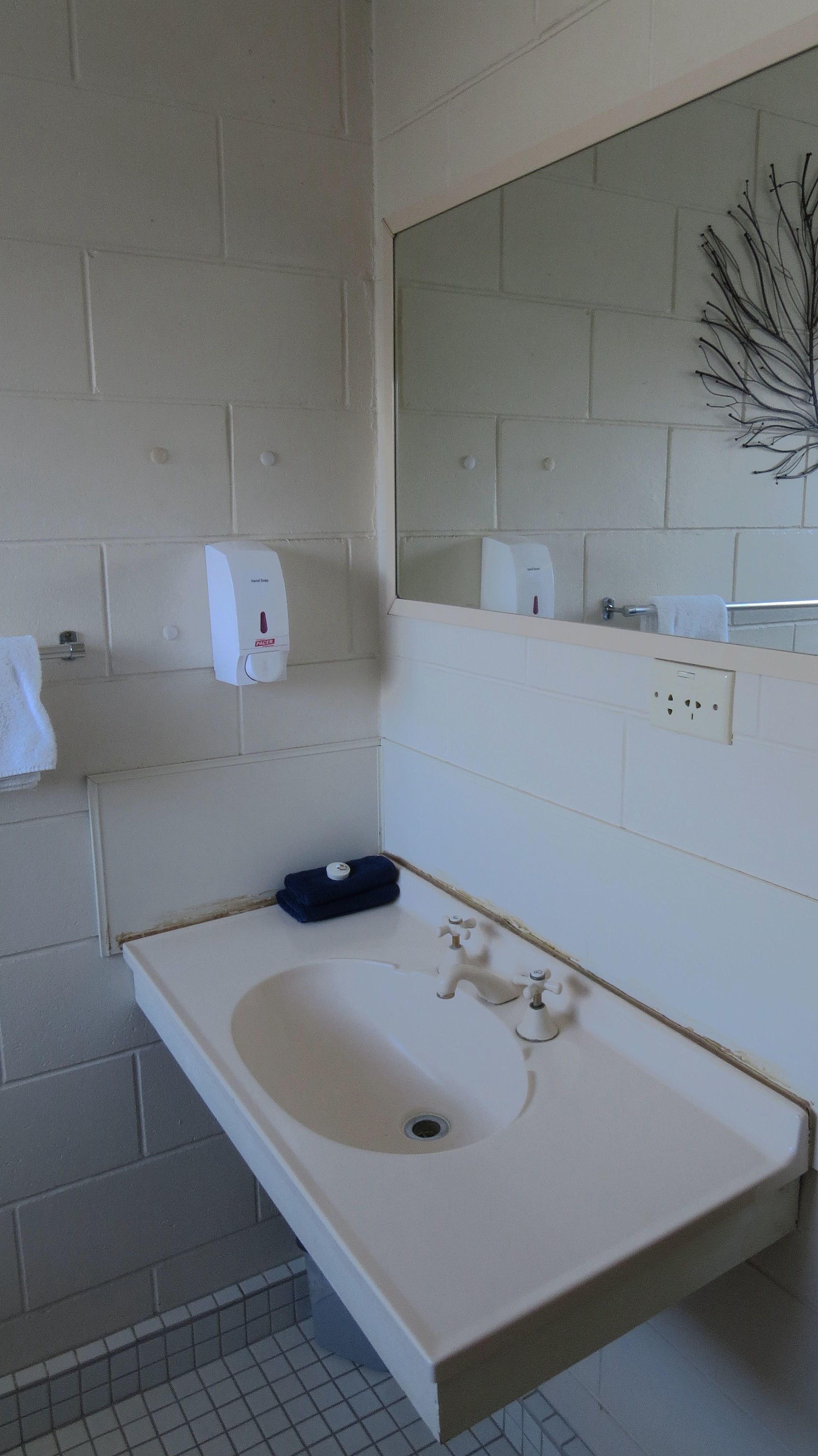Ein Badezimmer mit einem weißen Waschbecken, zwei Wasserhähnen, einer Seifenspender, einem Handtuchhalter und einem Spiegel.