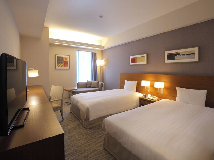 A hotel room with two beds, a desk, a chair, a TV, and a lamp.
