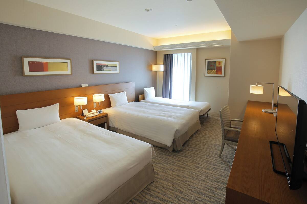 Ein Hotelzimmer mit zwei Betten, einem Schreibtisch mit Computer und einem Fernseher. An den Wänden sind Lampen befestigt und es gibt ein Fenster mit Vorhängen.
