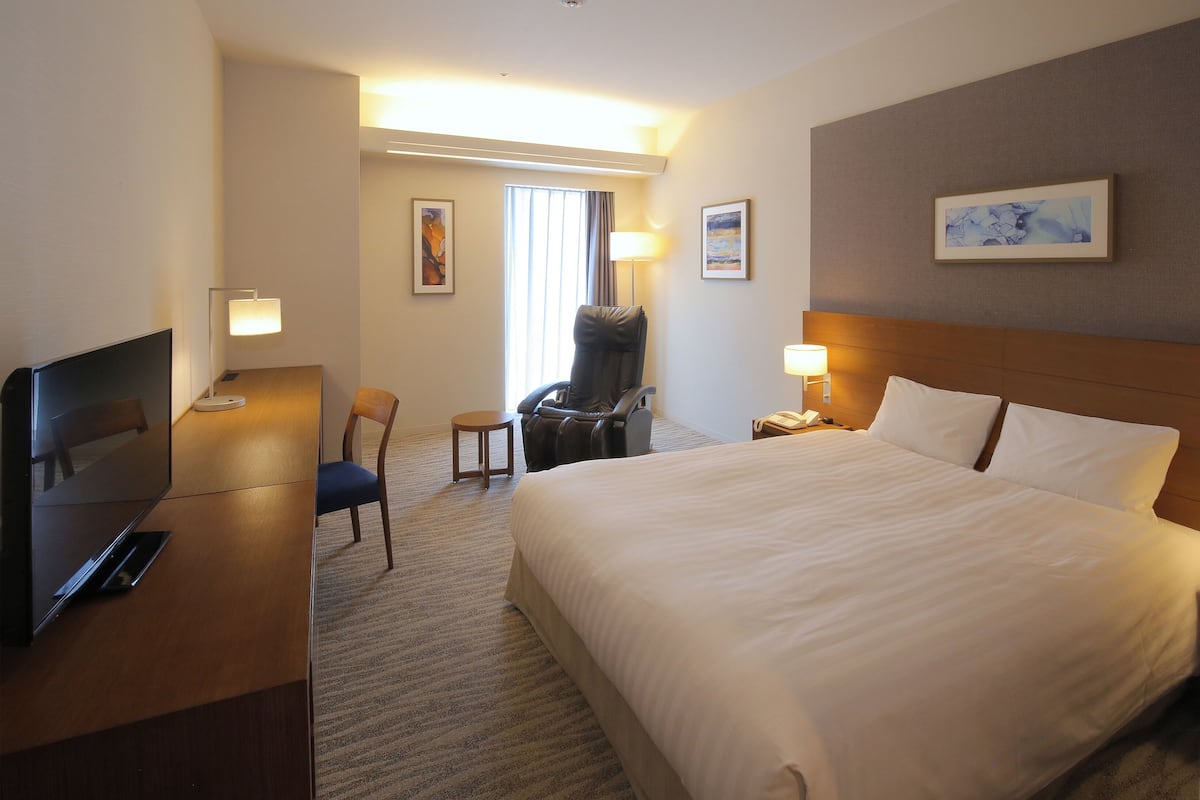 Ein Hotelzimmer mit einem großen Bett, einem Schreibtisch mit Fernseher, einem Sessel und einer Schreibtischlampe.