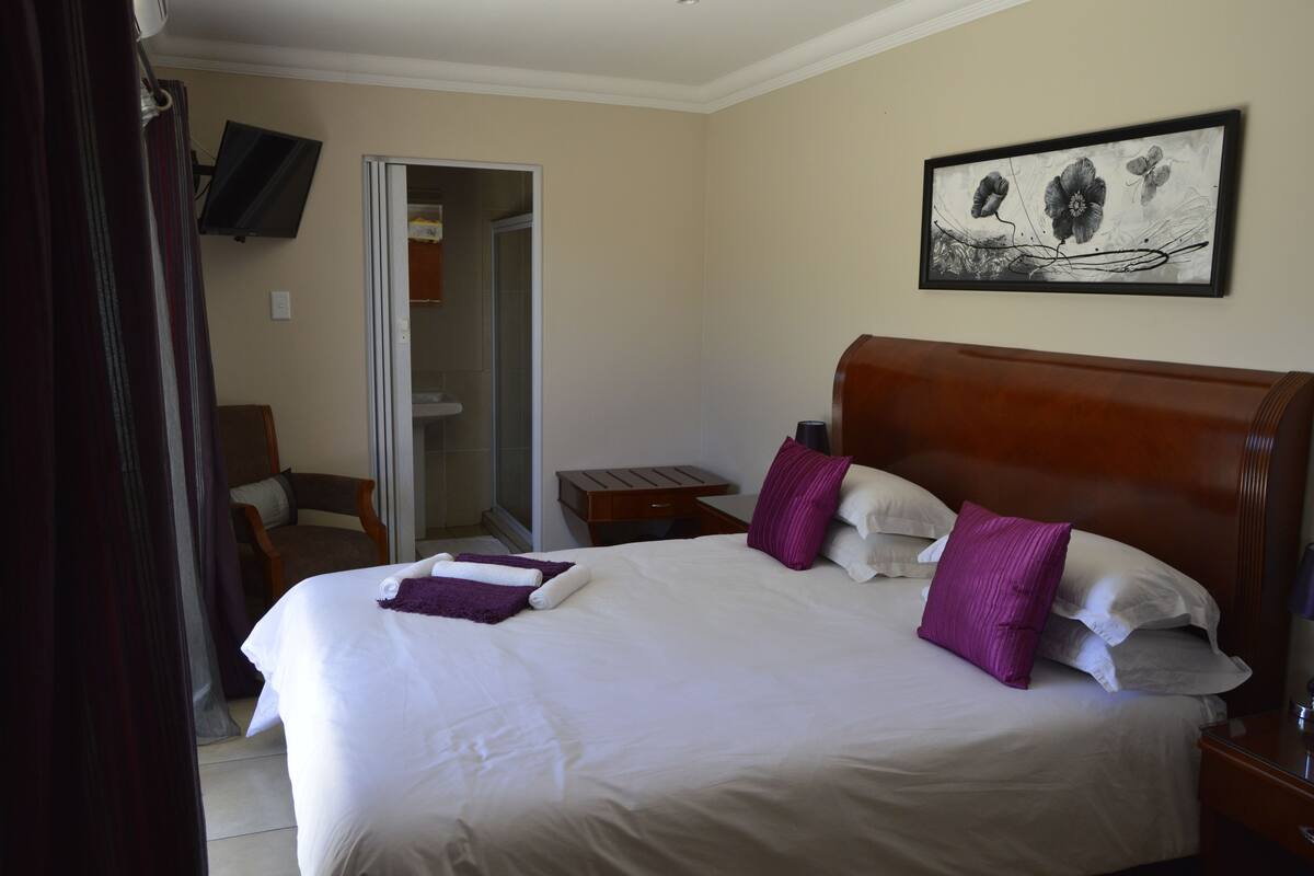 Habitación de hotel con una cama grande, cabecero de madera, un televisor de pantalla plana montado en la pared y una obra de arte enmarcada sobre la cama.