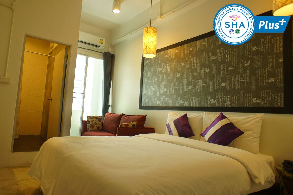 A hotel room with a large bed, a flat-screen TV, a wall-mounted air conditioning unit, and a window with curtains.