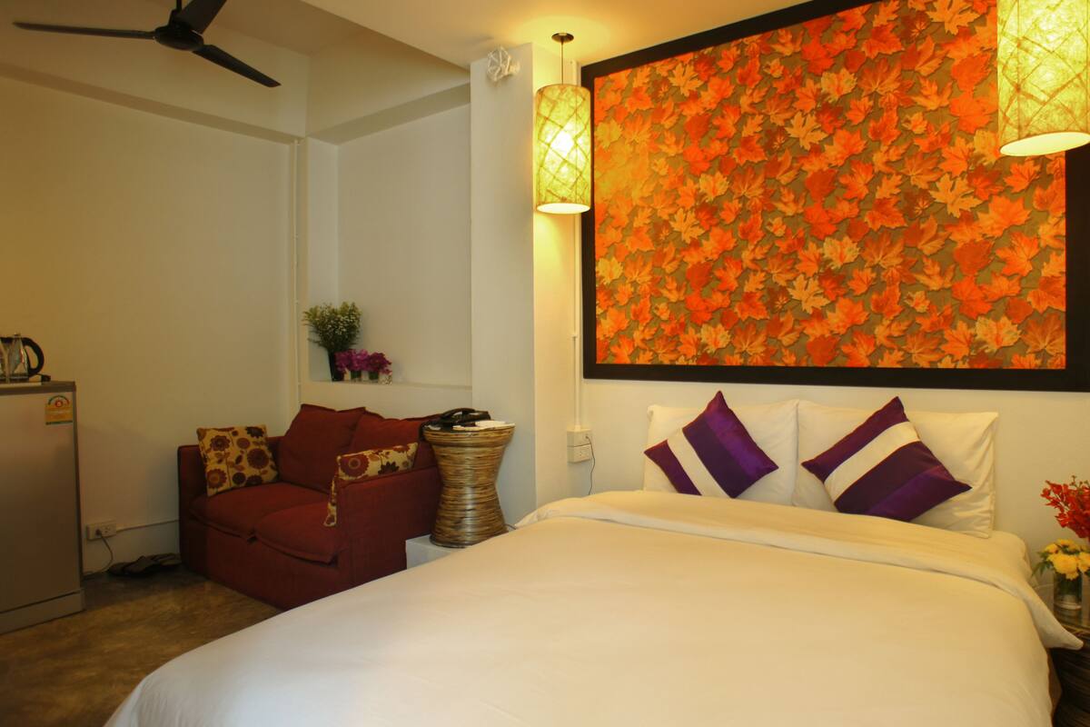 A hotel room with a bed, a red armchair, a small table with a vase of flowers, and a wall with a floral wallpaper.