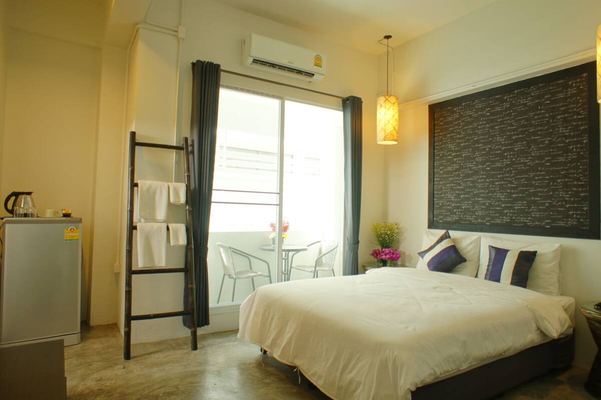 A hotel room with a bed, a desk, a chair, a refrigerator, a window with curtains, and a wall with a chalkboard.