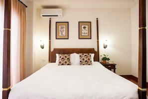 A neatly made bed with a wooden headboard, two wall-mounted air conditioning units, and two framed botanical prints on the wall.