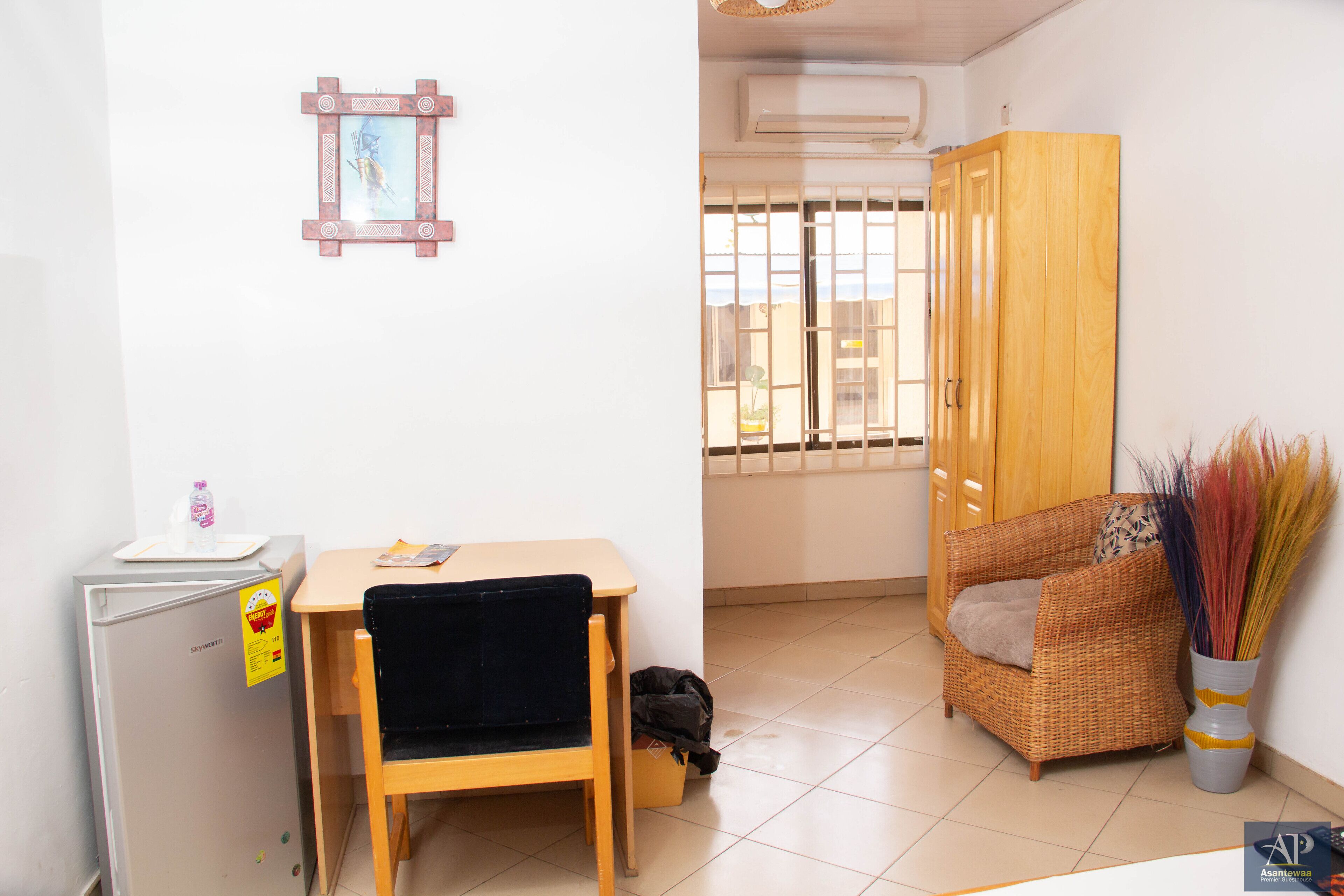 A compact room with a small table, a chair, a refrigerator, a wicker sofa, and a window with bars.