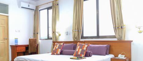 Una cama bien hecha con ropa de cama blanca y morada, una mesita de noche con un teléfono y una ventana con cortinas.