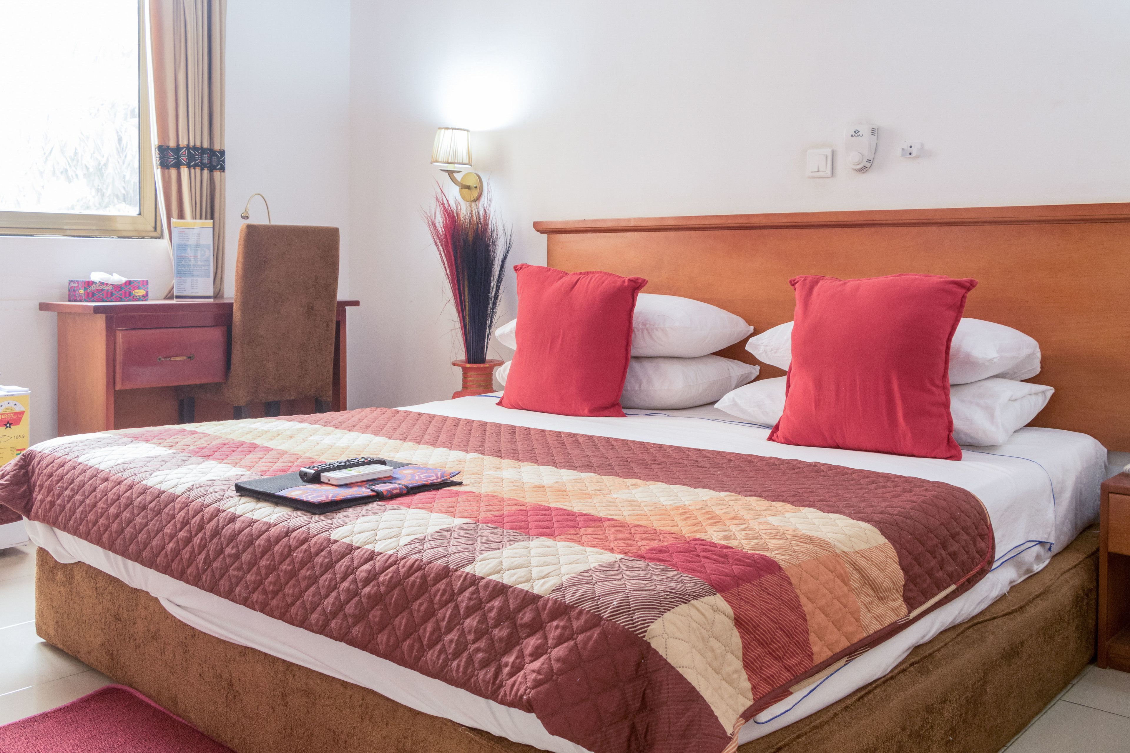 Een netjes opgemaakt bed met een gewatteerde spreideken en twee rode kussens, een nachtkastje met een lamp en een raam met gordijnen.
