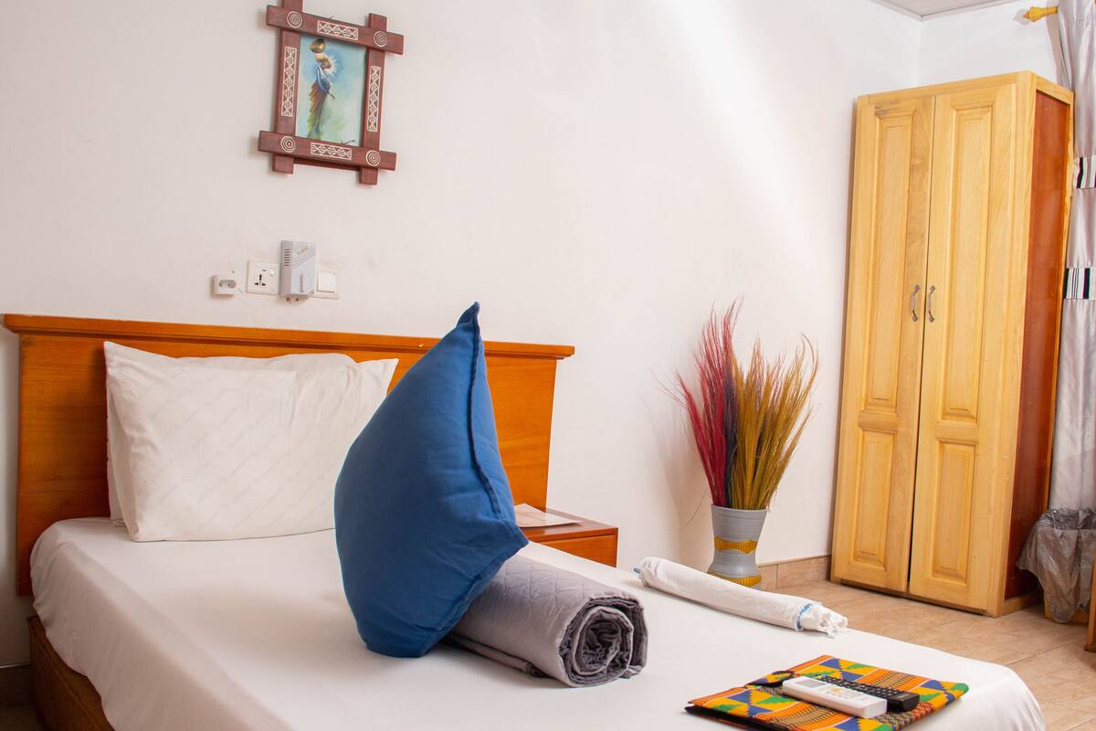 A neatly made bed with a blue pillow and a rolled-up towel, a bedside table with a remote control, a vase with dried plants, and a wooden wardrobe.