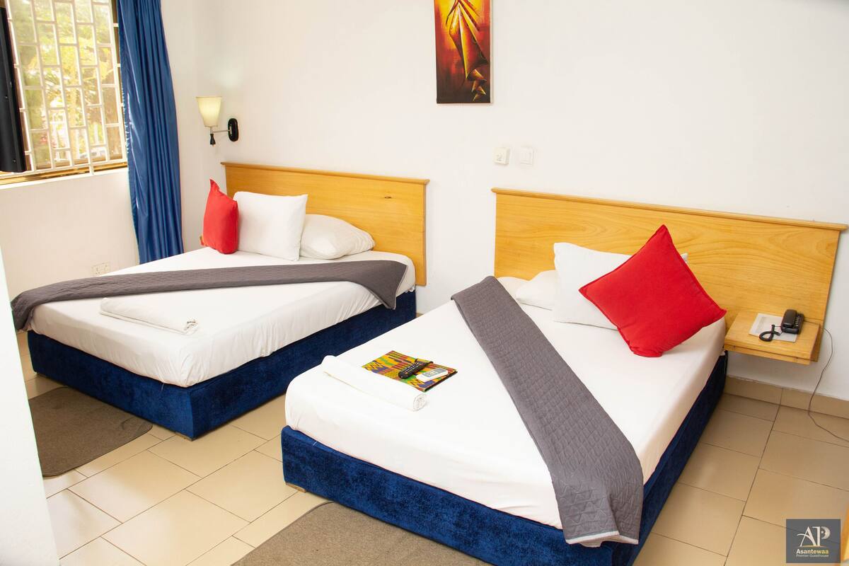 A hotel room with two beds, each with a wooden headboard, white bedding, and red and gray pillows. There is a small bedside table with a lamp and a phone.