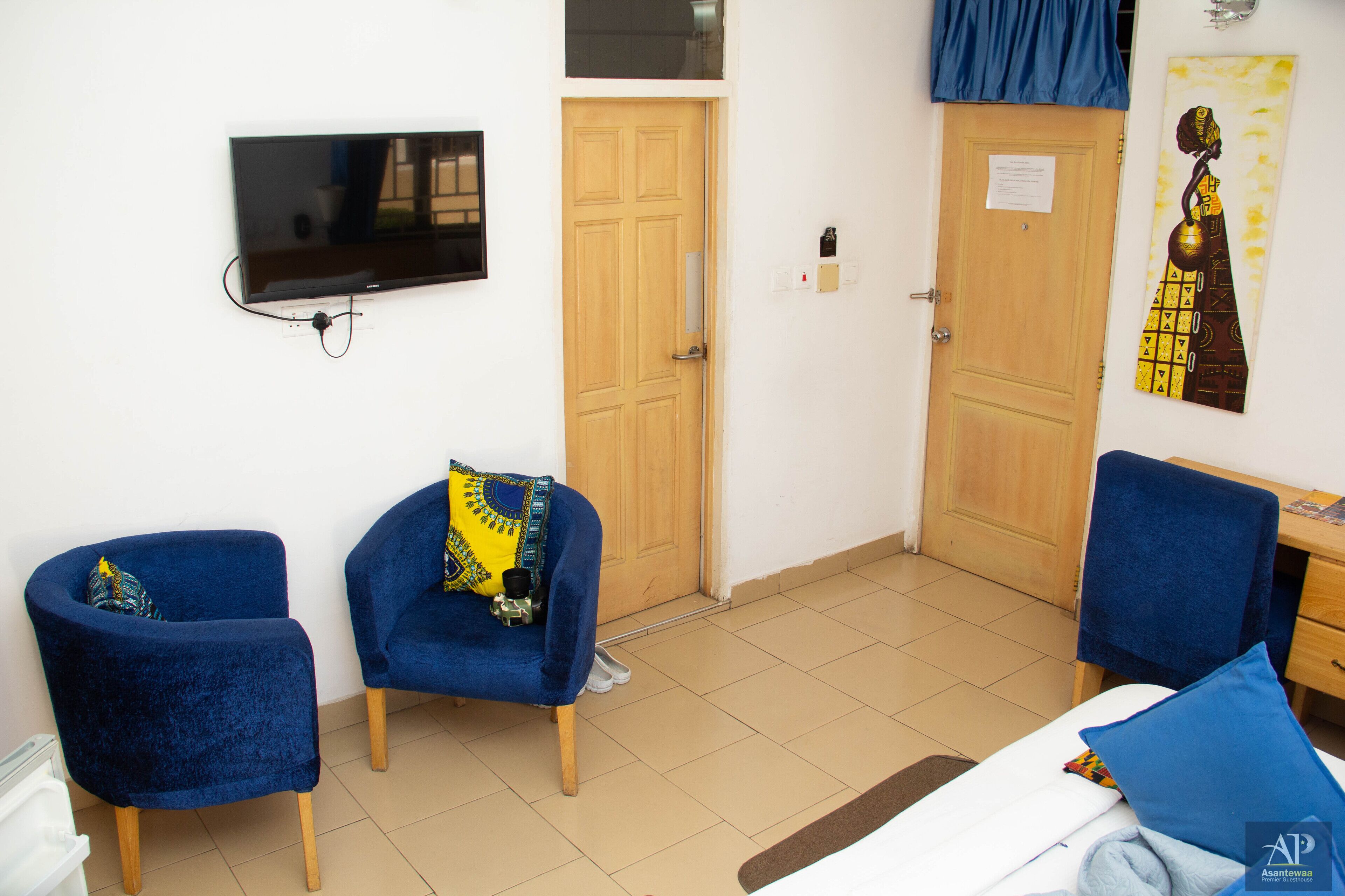 A hotel room with a bed, two blue armchairs, a television mounted on the wall, and two wooden doors.