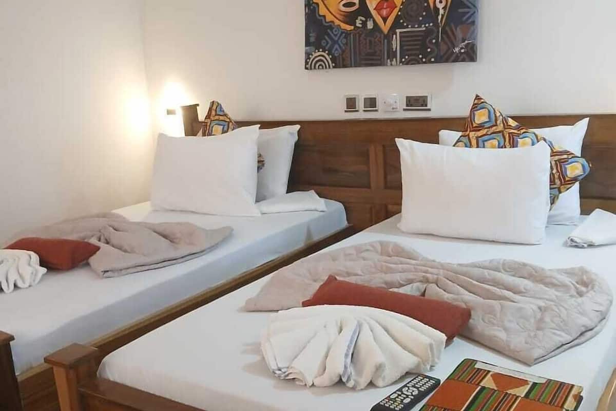 A hotel room with two single beds, a wooden headboard, a painting above the beds, and a bedside table with a lamp.