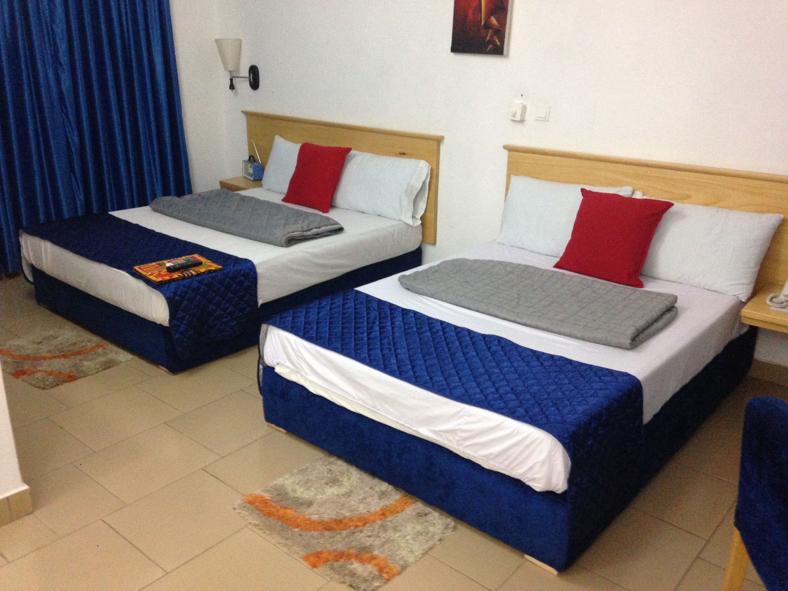 A hotel room with two beds, each with blue bedding and red pillows. There is a nightstand with a lamp and a small table with a phone. The room has a tiled floor and a rug with orange and gray patterns.