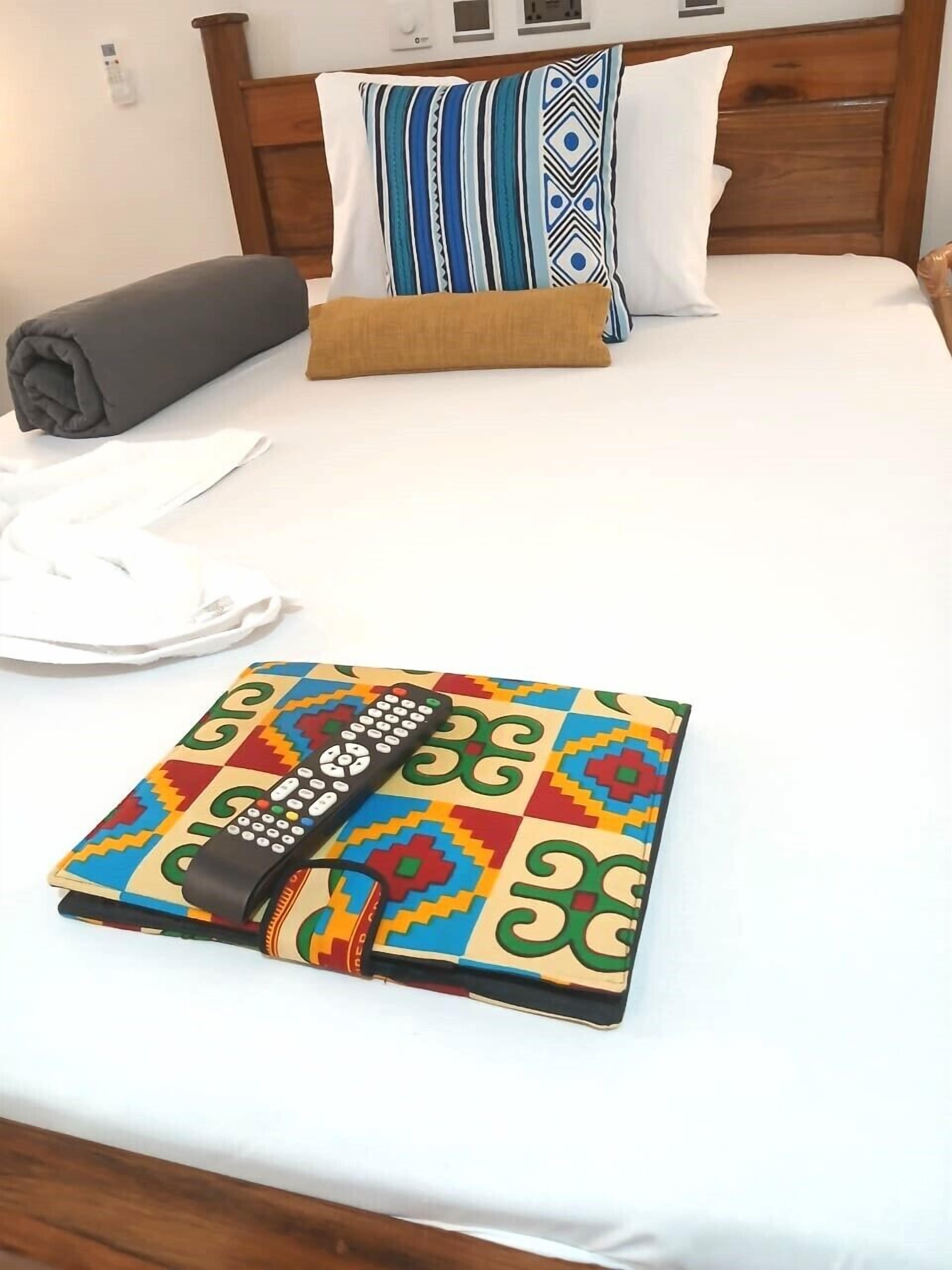 Ein ordentlich bezogenes Bett mit einer gemusterten Decke, Kissen und einer Fernbedienung auf einer farbenfrohen Matte.