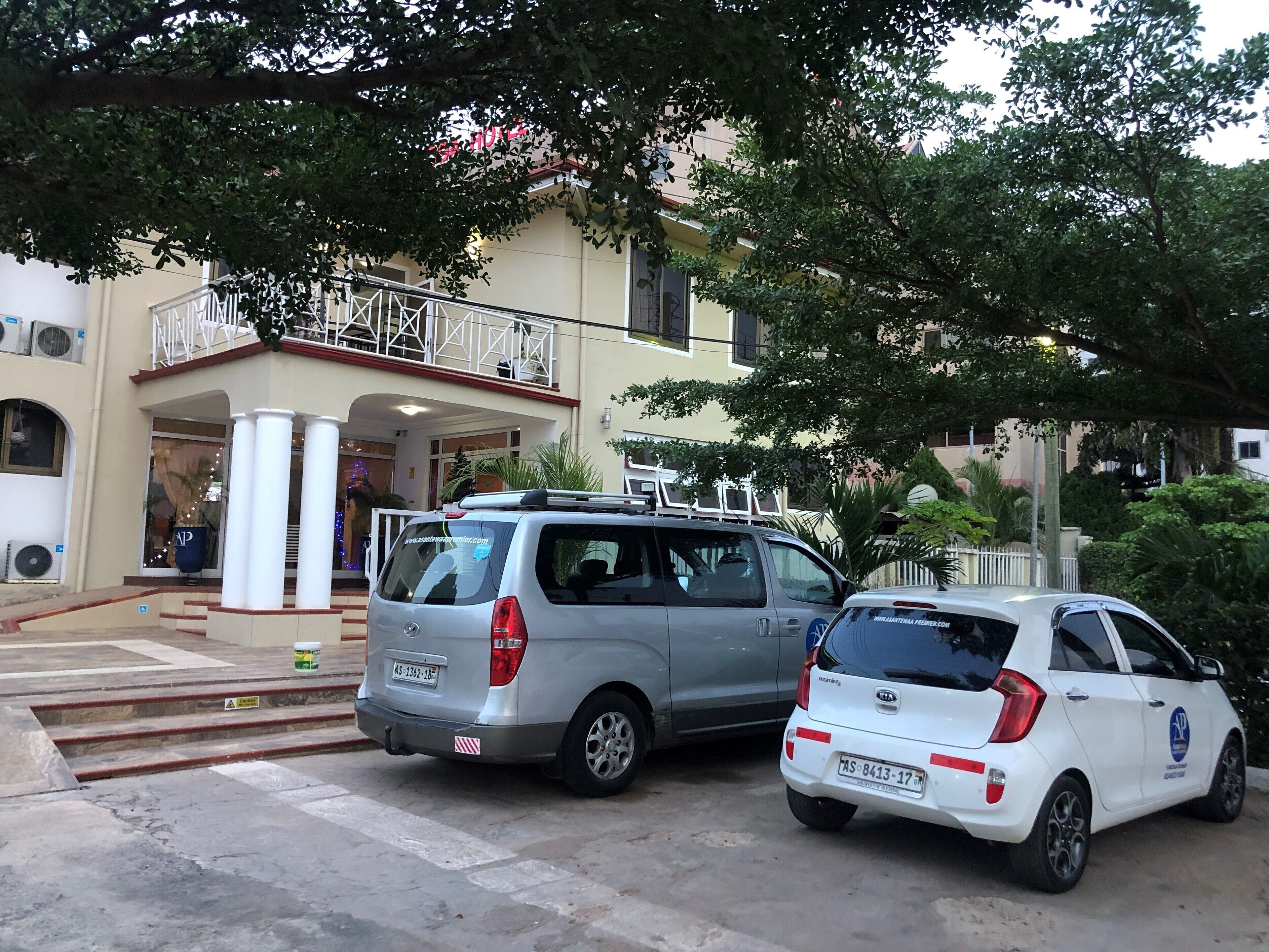 A hotel entrance with two parked cars, a balcony, and a staircase leading to the entrance.