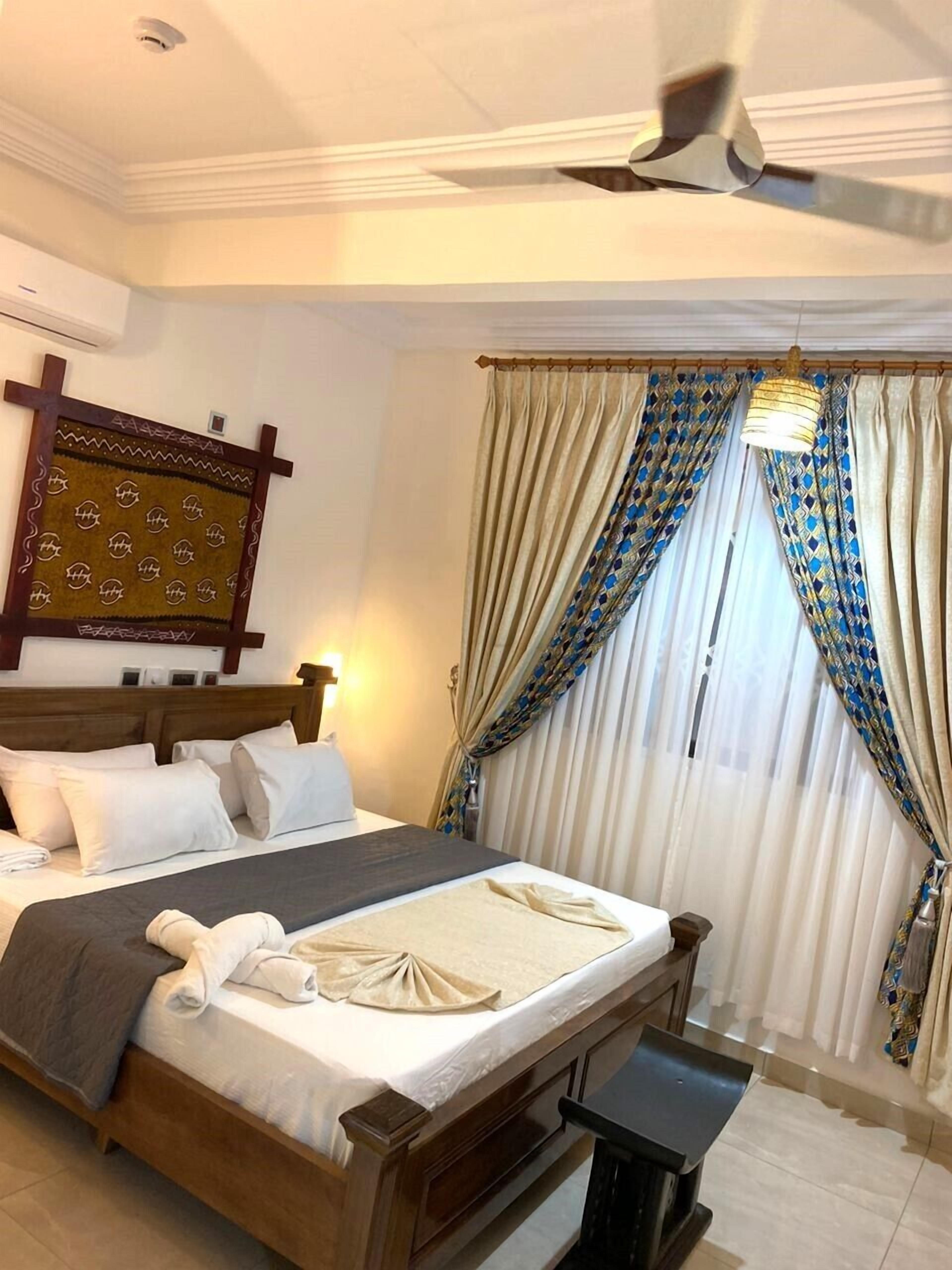 Habitación de hotel con cama de madera, mesita de noche, ventilador de techo y ventana con cortinas estampadas.