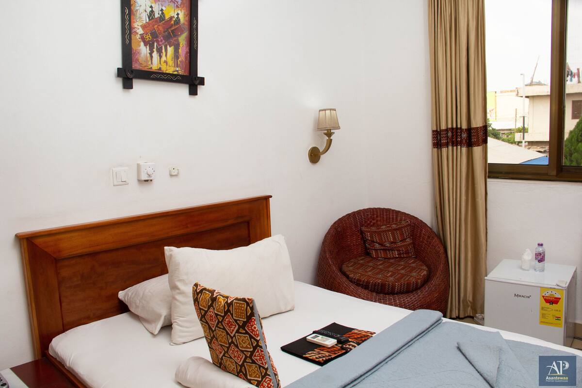 A hotel room with a wooden headboard, a bed with white and patterned pillows, a wicker chair, a bedside table with a bottle, and a wall-mounted lamp.