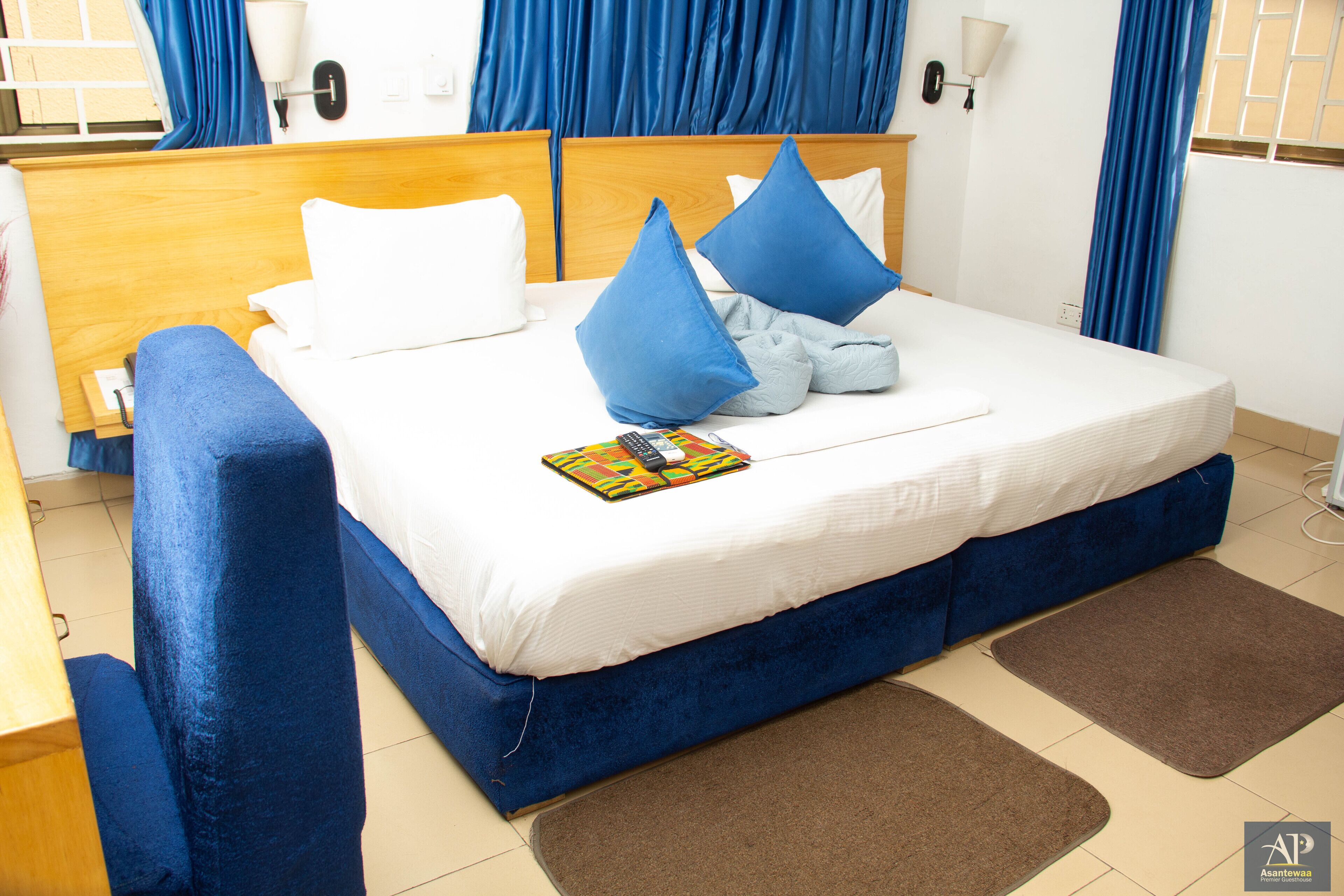A hotel room with a bed, blue curtains, a wooden headboard, and a brown floor mat.