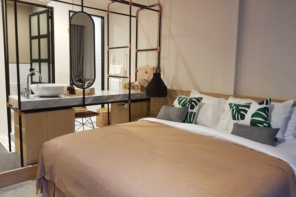 Una habitación de hotel moderna con una cama grande, un baño con un espejo amplio y una lámpara colgante.