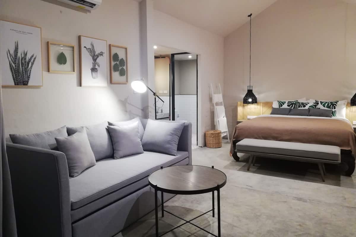 Un dormitorio moderno con un sofá gris, una mesita redonda pequeña, una cama con cabecera marrón y aire acondicionado instalado en la pared.