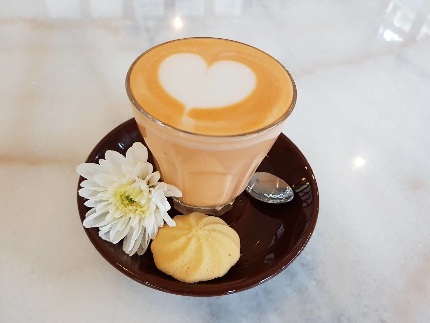 A glass of coffee with a heart design on the foam, a white flower, and a cookie on a dark plate.