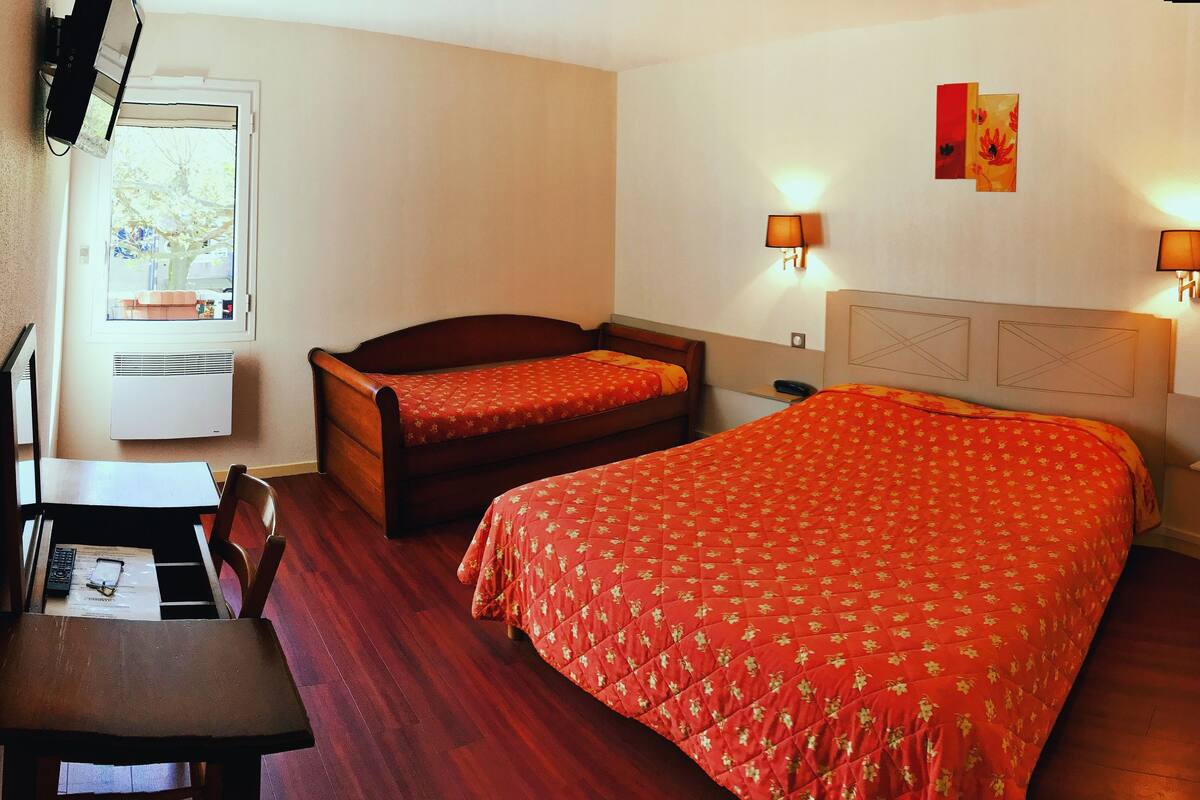 Ein Hotelzimmer mit Bett, Schreibtisch und Stuhl. Zwei Wandleuchten und ein Fenster mit Blick auf Bäume.