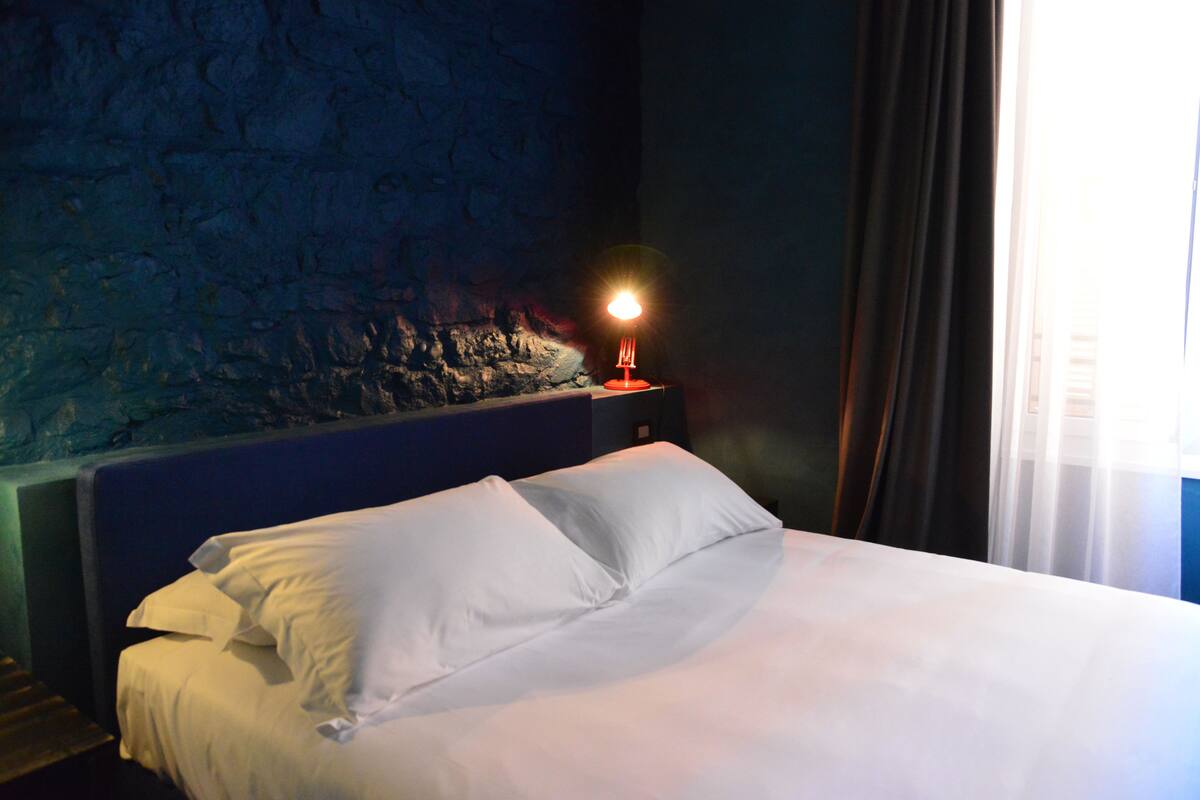 Una cama bien tendida con sábanas blancas en una habitación con pared de piedra y una mesita de noche con una lámpara.